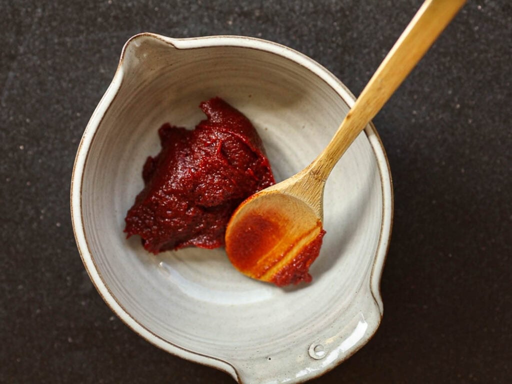 A ceramic bowl with a scoop of dark red gochujang paste is placed against a dark background. A wooden spoon rests in the bowl, partially dipped in the paste.